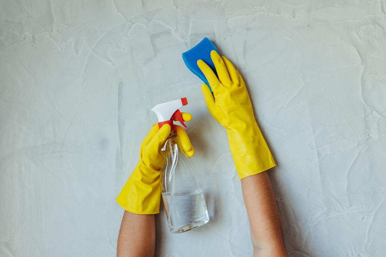 Gros plan sur des mains gantées de jaune qui nettoient un mur avec un vaporisateur et une éponge.